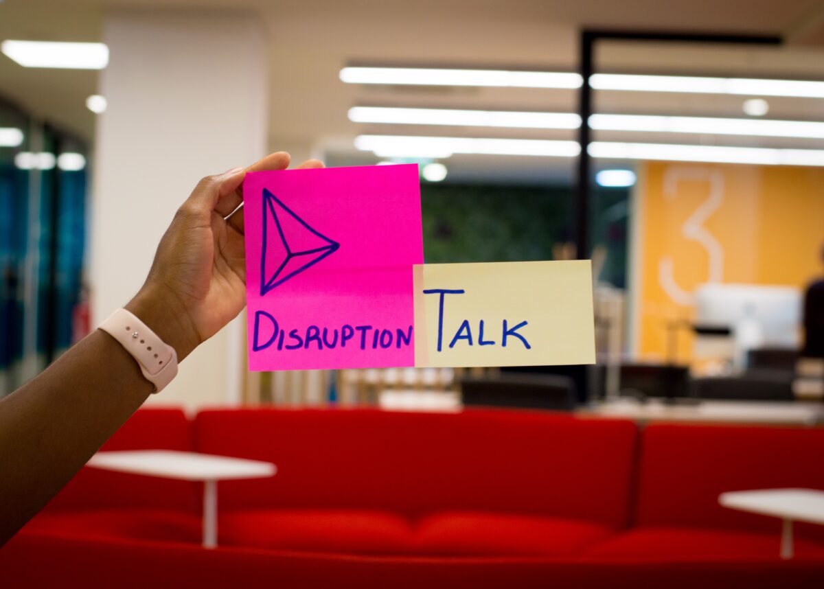 A hand places a post-it on an office window that reads Disruption-Talk
